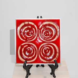 May include: Square ceramic tile with a glossy red background and four cream-colored spiral designs. The tile is framed in silver and displayed on a black easel. The artwork has a mid-century modern aesthetic.