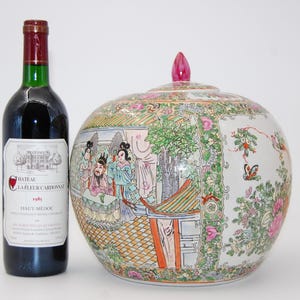 Pode incluir: Um pote de cerâmica redondo com tampa e remate rosa, decorado com cenas pintadas à mão de figuras num jardim e padrões florais. Ao lado, encontra-se uma garrafa de vinho de vidro escuro com um rótulo que diz Chateau La Fleur Cardonnat 1985.