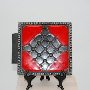 Könnte beinhalten: Quadratische dekorative Fliese mit rotem und silbernem Design. Die Fliese hat ein strukturiertes, geometrisches Muster und ist von einem silbernen Rahmen eingefasst. Sie wird auf einem schwarzen Metallständer präsentiert.
