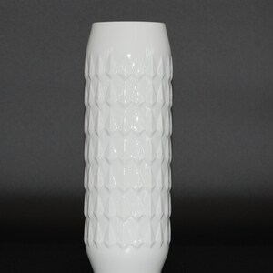 Könnte beinhalten: Eine hohe, zylindrische weiße Vase mit einem sich wiederholenden geometrischen Muster aus erhabenen, rautenförmigen Facetten auf der Außenfläche. Die Vase hat eine glatte, glänzende Oberfläche und einen einfachen, runden oberen Rand.