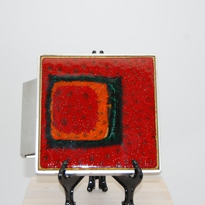 May include: Square ceramic tile with a textured red surface, featuring a central square design in orange and green. The tile is framed in a silver metal border and displayed on a black stand.