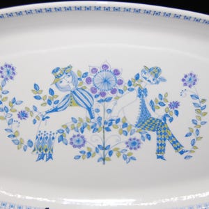 May include: A white rectangular ceramic serving platter featuring a blue and green illustration of two figures sitting among stylized flowers and foliage. The rim of the platter is decorated with a repeating blue geometric pattern along the edge.