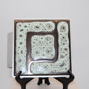 May include: Square ceramic tile with a geometric design. The tile features a light green speckled background with a dark brown square and border. The tile is framed in a silver metal. The tile is displayed on a black stand.
