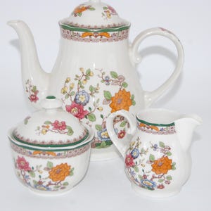 Könnte beinhalten: Ein weißes Porzellan-Teeservice mit floralen Mustern. Das Set beinhaltet eine Teekanne, eine Zuckerdose mit Deckel und einen Sahnekännchen. Das florale Design zeigt orange, rosa, blaue und gelbe Blumen mit grünen Akzenten.
