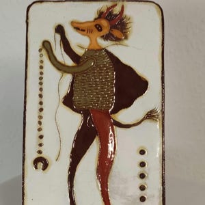 Könnte beinhalten: Rechteckige Keramikfliese mit einer stilisierten Figur mit hirschähnlichem Kopf, die einen Umhang trägt und eine Perlenkette hält. Die Figur ist in Braun, Rot und Beige vor einem weißen Hintergrund mit braunem Rand dargestellt.