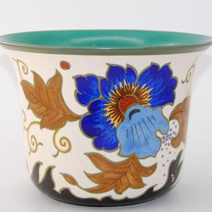 Può includere: Un vaso per piante in ceramica con base color crema decorato con un grande fiore blu vivace dal centro giallo, circondato da foglie marroni e viticci dorati. Il bordo interno è verde acqua tinta unita e la base ha un bordo nero smerlato.