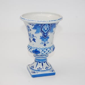 Könnte beinhalten: Eine weiße, urnenförmige Keramikvase mit komplexen blauen Blumen- und geometrischen Mustern. Das Gefäß hat einen breiten, ausgestellten Rand, einen bauchigen Mittelteil mit Gitterdesign und einen quadratischen Sockel, alles mit blauen Motiven.