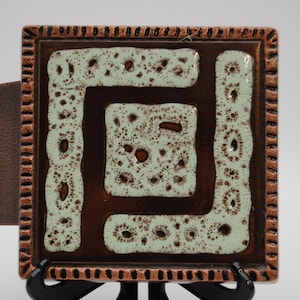 May include: Square ceramic tile with a brown and light green geometric design. The tile has a textured border and a dark brown backing. The tile is displayed on a black stand against a white background.