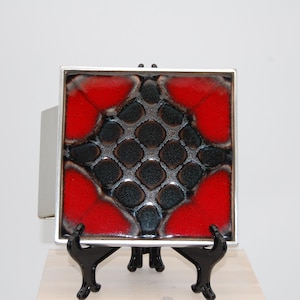 Könnte beinhalten: Quadratische Keramikfliese mit einem geometrischen Design in Rot und Schwarz, eingefasst in einem silbernen Rahmen. Die Fliese wird auf einem schwarzen Ständer präsentiert und zeigt ihre leuchtenden Farben und ihre strukturierte Oberfläche. Ein dekoratives Stück für die Inneneinrichtung.