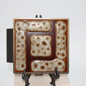 Puede incluir: Azulejo cerámico cuadrado con un diseño abstracto marrón y beige. El azulejo tiene una superficie texturizada en relieve con un borde marrón oscuro y un soporte de montaje negro en la parte posterior. El azulejo se exhibe sobre un soporte negro.