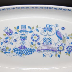 May include: A white rectangular ceramic serving platter featuring a blue, purple, and green line-art illustration of two figures sitting among flowers and a bird. The border of the platter is decorated with a repeating circular pattern in blue.