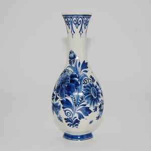 Op de afbeelding: Een witte keramische vaas met een bolvormige body en een slanke hals. De vaas is versierd met een blauw bloemmotief, met grote bloemen en ingewikkelde bladmotieven. De basis en rand hebben blauwe accenten.