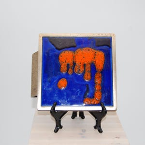 Puede incluir: Azulejo de cerámica cuadrado con fondo azul y diseño abstracto naranja. El azulejo está enmarcado en plata y se exhibe sobre un caballete negro. La obra de arte tiene una estética moderna de mediados de siglo.