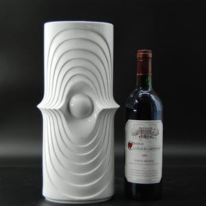 Könnte beinhalten: Eine weiße zylindrische Keramikvase mit einem Reliefmuster aus konzentrischen ovalen Ringen und einer zentralen Kugel, neben einer dunklen Weinflasche aus Glas mit dem Etikett Chateau La Fleur Cardonne 1981 Haut-Medoc.
