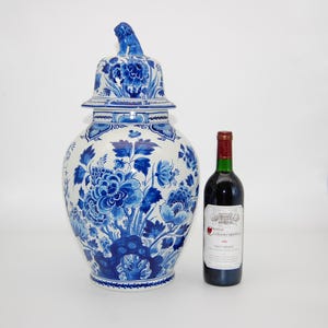 May include: A large white ceramic ginger jar with blue floral patterns and a lid topped with a lion figurine. The jar is positioned next to a dark glass wine bottle with a label reading Chateau La Fleur Cardonnat 1981 Haut-Medoc.