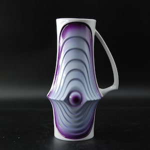 Könnte beinhalten: Ein weißer Keramikkrug mit einem markanten, erhabenen Mittelteil, der konzentrische, wellenförmige Rippen aufweist. Das Design ist mit einem tiefvioletten Farbverlauf akzentuiert, der in einen helleren Lavendelton übergeht.