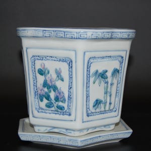 Könnte beinhalten: Weißer Keramik-Blumentopf mit blauen Blumen- und Bambusmotiven. Der quadratische Pflanztopf hat eine passende quadratische Untertasse. Das Design des Pflanztopfs umfasst blaue Verzierungen und einen dekorativen Rand.