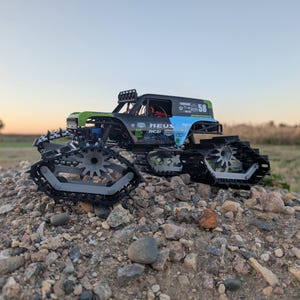 May include: A black, green, and blue RC truck with tank treads, parked on a pile of rocks. The truck has the number 58 on the side and the text "HEUS RCZ". The sky is a gradient of orange and blue.