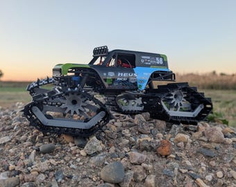 May include: A black, green, and blue RC truck with tank treads, parked on a pile of rocks. The truck has the number 58 on the side and the text "HEUS RCZ". The sky is a gradient of orange and blue.