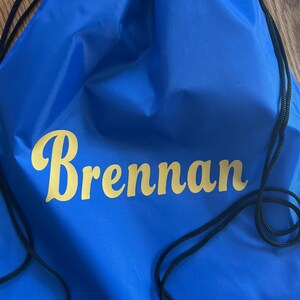 May include: A royal blue drawstring backpack with the name "Brennan" printed in a gold, cursive font. The bag has black drawstrings and is made of a smooth, possibly nylon, material.