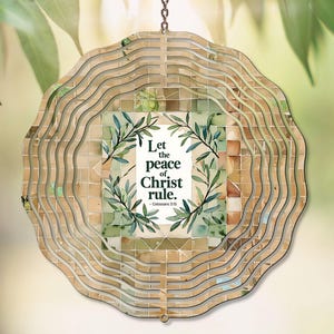 Könnte beinhalten: Ein dekorativer, mehrschichtiger Windspinner mit einem zentralen Quadrat mit dem Text "Lasst den Frieden Christi herrschen" und Olivenzweig-Illustrationen. Der Windspinner hat ein geschichtetes, wellenförmiges Design in Beige- und Brauntönen.