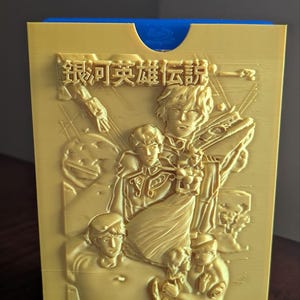 May include: A gold-colored 3D-printed card holder featuring a detailed relief of characters and Japanese text. The card holder has a blue interior and a rectangular shape. The embossed design includes figures and a spaceship.