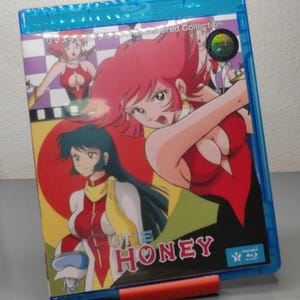 Op de afbeelding: Een blauwe Blu-ray-hoes met anime-illustraties van een vrouw met rood haar en een rode outfit. De titel "CUTIE HONEY" wordt weergegeven in een gestileerd lettertype. De hoes wordt vastgehouden door een rode standaard.