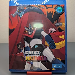 Puede incluir: Una caja azul de Blu-ray con el título "Great Mazinger". La portada presenta una ilustración dinámica del robot principal y otros personajes sobre un fondo rojo y naranja. El texto "Great Mazinger" es prominente. Fabricado en Japón.