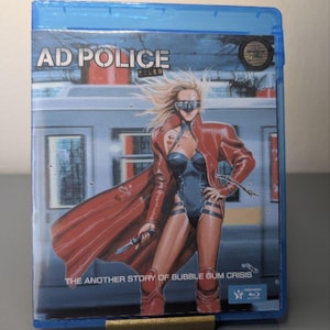 May include: A blue Blu-ray case with the title "AD POLICE FILES" and an illustration of a woman in a red coat and black bodysuit. The text "THE ANOTHER STORY OF BUBBLE GUM CRISIS" is also visible.