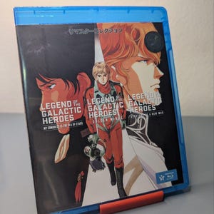 Puede incluir: Una caja Blu-ray azul con ilustraciones de "Legend of the Galactic Heroes". La portada muestra personajes en un tema de ópera espacial, con texto japonés y títulos en inglés como "My Conquest is the Sea of Stars" y "Golden Wings."