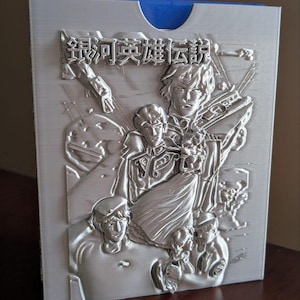 May include: A silver-colored, rectangular case with a blue interior. The front features a detailed relief of multiple figures and Japanese text. The artwork appears to be a scene from a science fiction or fantasy story.