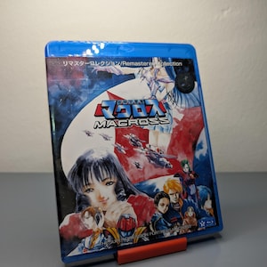 Puede incluir: Una caja azul de Blu-ray de la serie de anime "Macross". La portada muestra a una mujer con cabello negro, aviones de combate y otros personajes. El texto incluye "Macross" y caracteres japoneses.