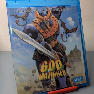 Puede incluir: Una caja de Blu-ray azul con el título "GOD MAZINGER" en letras coloridas. La portada muestra un gran robot con una espada, dragones y un castillo. La caja está sujeta por un soporte rojo. El texto "Remastered Collection" es visible.