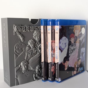 Puede incluir: Un estuche gris con ilustraciones en relieve y texto japonés, que contiene varias cajas de Blu-ray. Las cajas tienen lomos azules y presentan ilustraciones y texto en japonés e inglés, incluyendo "Legend of the Galactic Heroes."