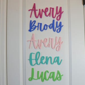 Puede incluir: Una puerta blanca con recortes de nombres de madera coloridos. Los nombres, en una fuente cursiva, son Avery (rosa), Brody (azul), Avery (rosa claro), Elena (turquesa) y Lucas (verde). La puerta tiene una manija negra.