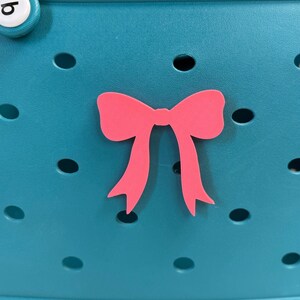 Puede incluir: Un zueco de espuma color verde azulado con un adorno en forma de lazo rosa brillante en la parte delantera. El zapato tiene una serie de orificios de ventilación circulares en la superficie, con dos botones blancos circulares en el borde superior, cada uno con una letra negra.