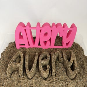 Puede incluir: Un letrero de color rosa brillante impreso en 3D con el nombre Avery en una fuente negrita sin remates, colocado sobre el nombre Avery formado directamente en un lecho de arena marrón, creando un contraste táctil entre el plástico y la arena.