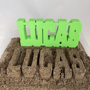 Puede incluir: Un letrero impreso en 3D de color verde lima brillante con el nombre LUCAS en una fuente negrita y cuadrada, colocado directamente sobre una impresión idéntica del nombre LUCAS presionada en un lecho de arena marrón granulada.