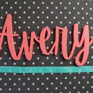 Puede incluir: Un recorte de nombre "Avery" en cursiva de color coral. El nombre mide aproximadamente 25 cm de largo, medido con una cinta m&eacute;trica turquesa. El fondo es una tela gris oscuro con lunares blancos.