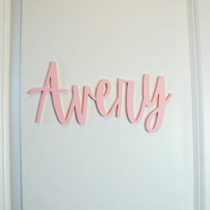 Puede incluir: Un letrero de madera rosa con el nombre "Avery" en escritura cursiva est&aacute; fijado a una pared blanca. El letrero es de un tono rosa claro, con las letras unidas. La pared tiene paneles verticales.