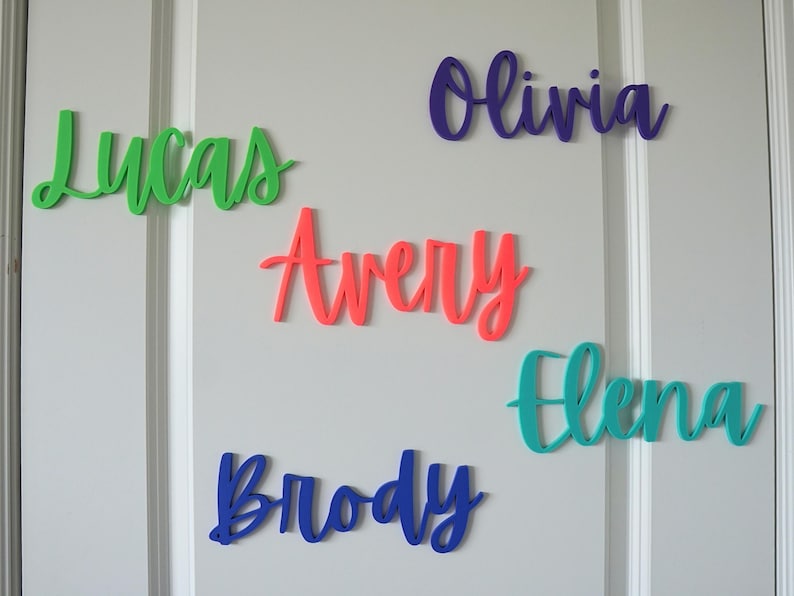 Puede incluir: Letreros de nombres de madera coloridos en verde, morado, azul y coral. Los nombres "Lucas", "Olivia", "Avery", "Brody" y "Elena" se muestran en una fuente cursiva, perfectos para la decoraci&oacute;n de la guarder&iacute;a o regalos personalizados.