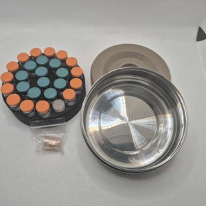 May include: A stainless steel container with a lid, next to a circular black holder with small vials. The vials have orange and teal caps. A small bag of capsules is also present.
