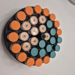 May include: A circular black holder with multiple small glass vials. The vials have orange and teal caps. The holder is designed to organize and display the vials. The vials are arranged in a circular pattern.