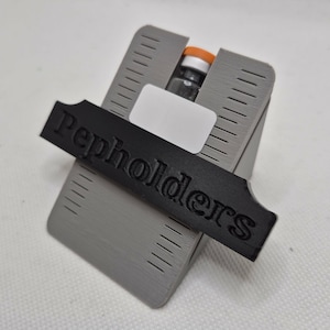 May include: A gray 3D-printed holder with a black label that reads "Pepholders." The holder is designed to cradle a small glass vial with an orange cap. The holder has a rectangular shape with angled sides and vertical lines.