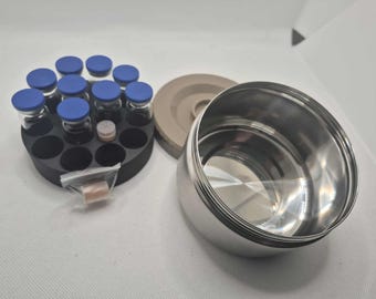 May include: A collection of laboratory equipment including a stainless steel container, a black circular rack holding vials with blue caps, and a beige disc. A small bag with a brown object is also present.