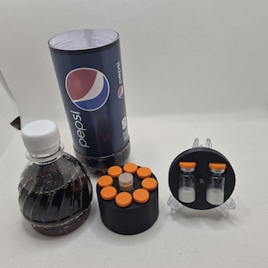 May include: A clear plastic bottle with a white cap, a Pepsi can, a black cylinder with orange caps, and two small vials on a clear stand. The Pepsi can is dark blue with the Pepsi logo. The cylinder has a central beige cap.