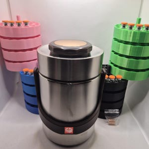 May include: A stainless steel insulated food container with a black handle and lid, surrounded by several colorful, tiered holders. The holders are pink, blue, green, and black, each holding small vials with orange caps.
