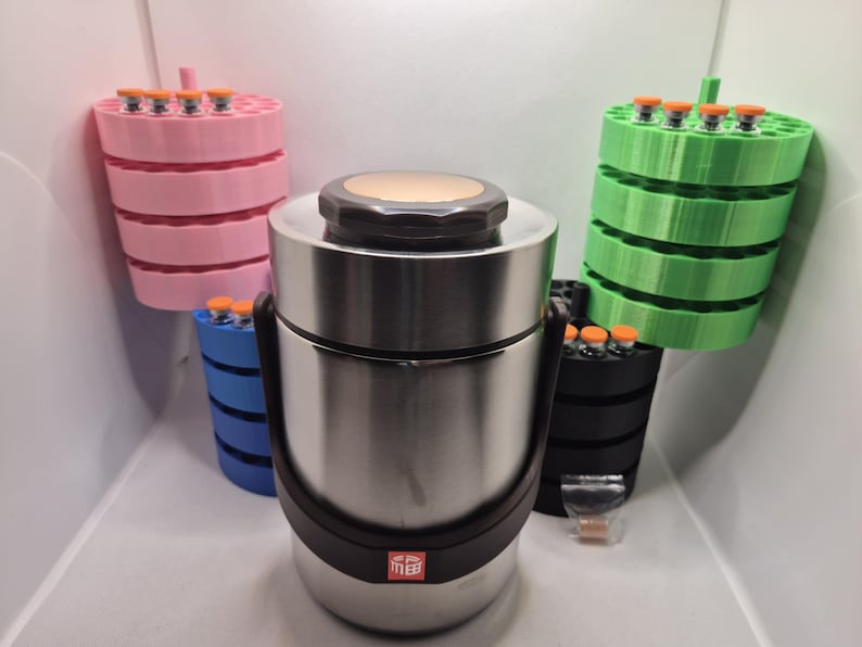 May include: A stainless steel insulated food container with a black handle and lid, surrounded by several colorful, tiered holders. The holders are pink, blue, green, and black, each holding small vials with orange caps.