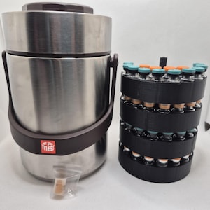 May include: A stainless steel insulated container with a brown handle and lid, next to a black cylindrical rack holding multiple small glass vials with teal and orange caps. The container has a brown band and a red square logo.