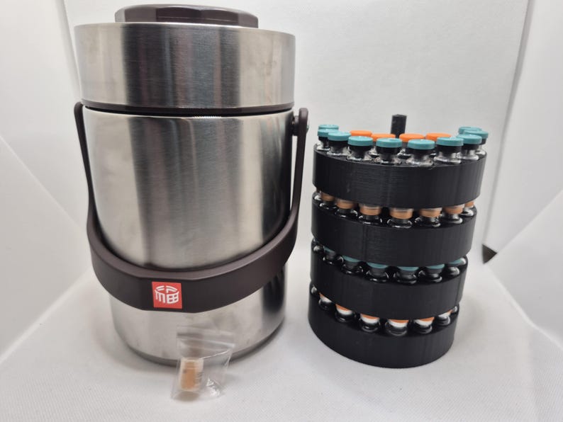 May include: A stainless steel insulated container with a brown handle and lid, next to a black cylindrical rack holding multiple small glass vials with teal and orange caps. The container has a brown band and a red square logo.